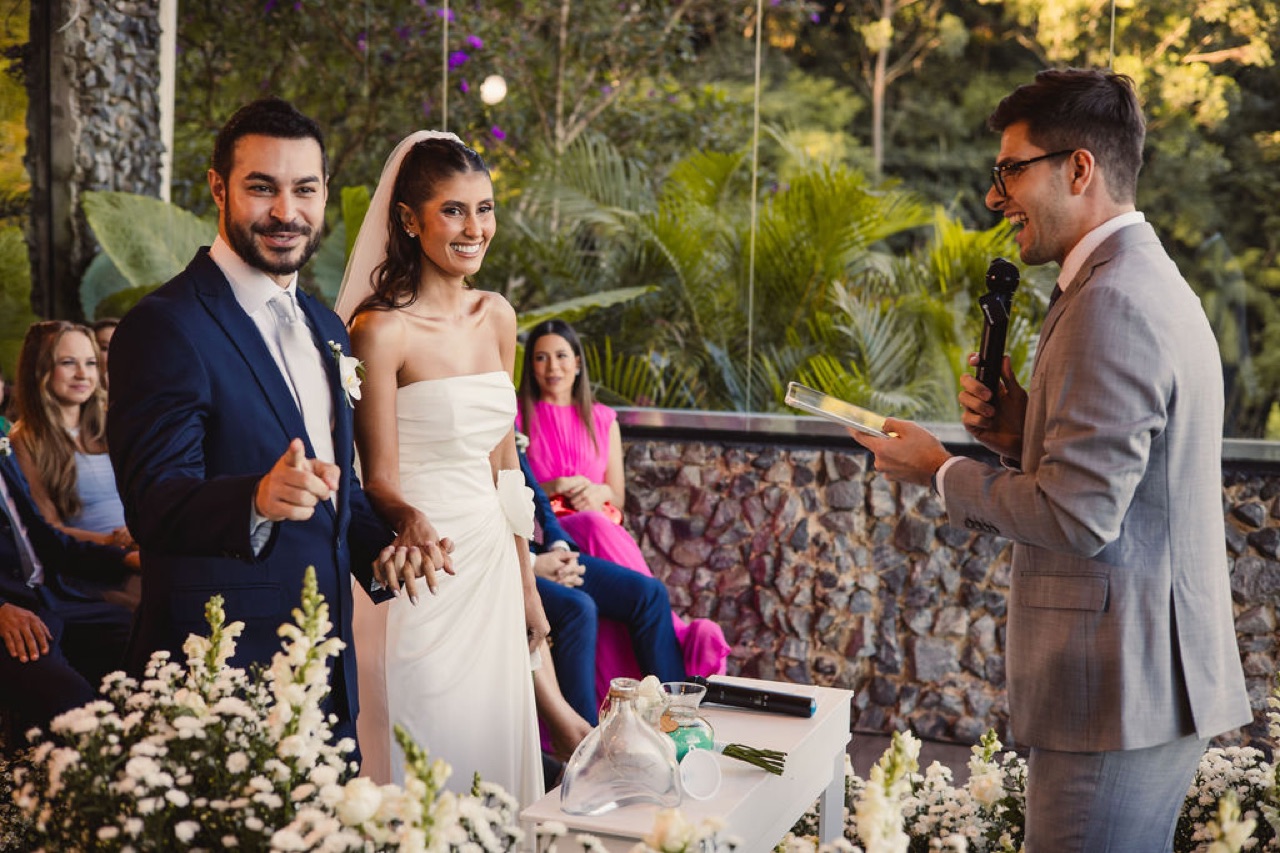 Noivos sorriem mostrando as alianças diante do celebrante, em cerimônia ao ar livre com flores brancas e muro de pedra