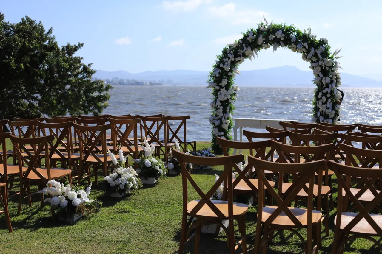 Cerimônia à beira da água no Indaiá em Florianópolis: arco de flores brancas diante da água e das montanhas, com fileiras de cadeiras de madeira sobre a grama.