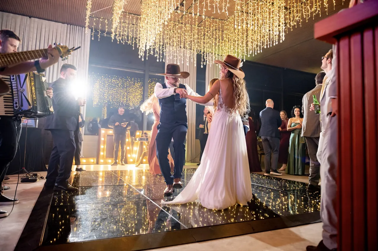 Casal dança de mãos dadas, com a banda ao lado e luzes douradas sobre a pista.