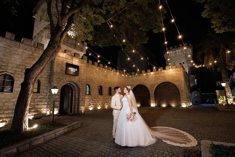 Noivos se beijam no pátio de pedra diante do castelo do Indaiá em Blumenau, à noite, sob cordões de luzes.