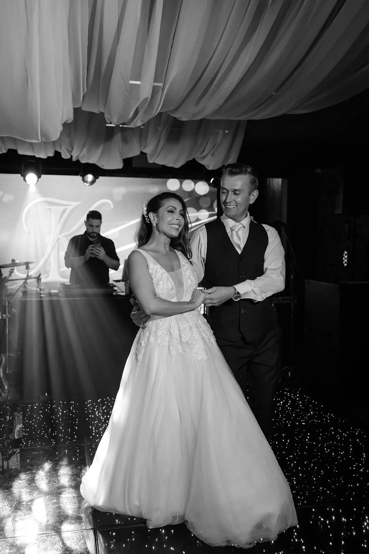 O casal sorrindo junto na pista de dança, sob tecidos brancos e luzes da festa, em uma fotografia em preto e branco.