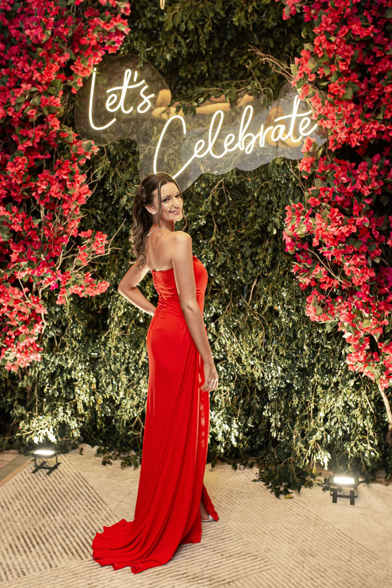 Formanda de vestido vermelho diante do painel floral iluminado da celebração.