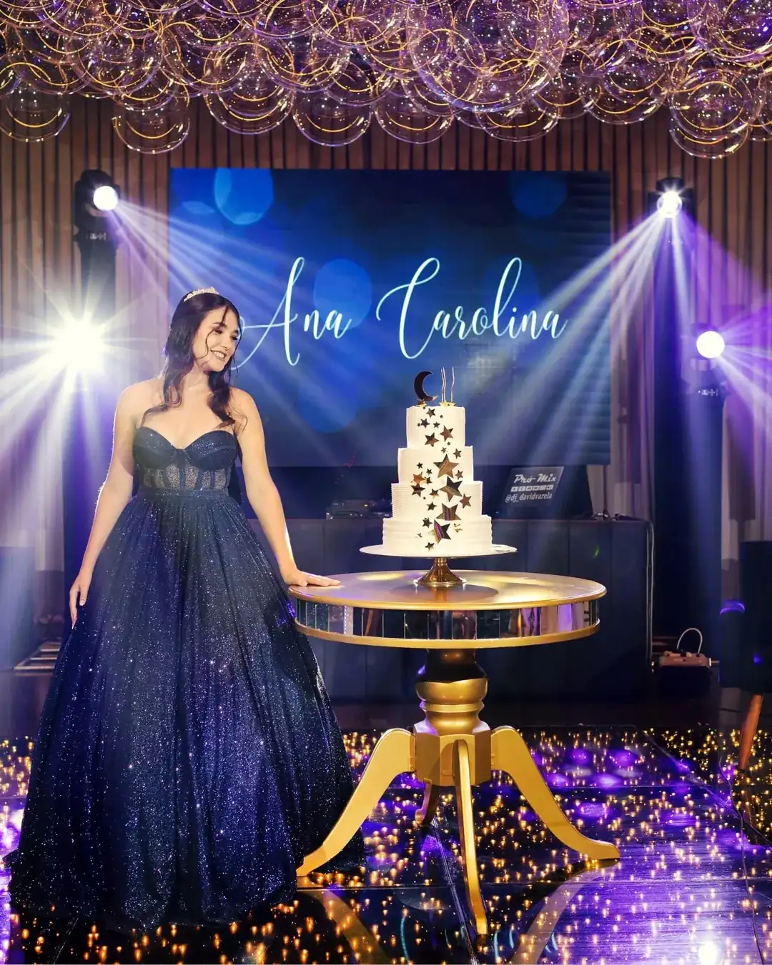 Debutante junto ao bolo decorado com estrelas, sob as luzes da festa.