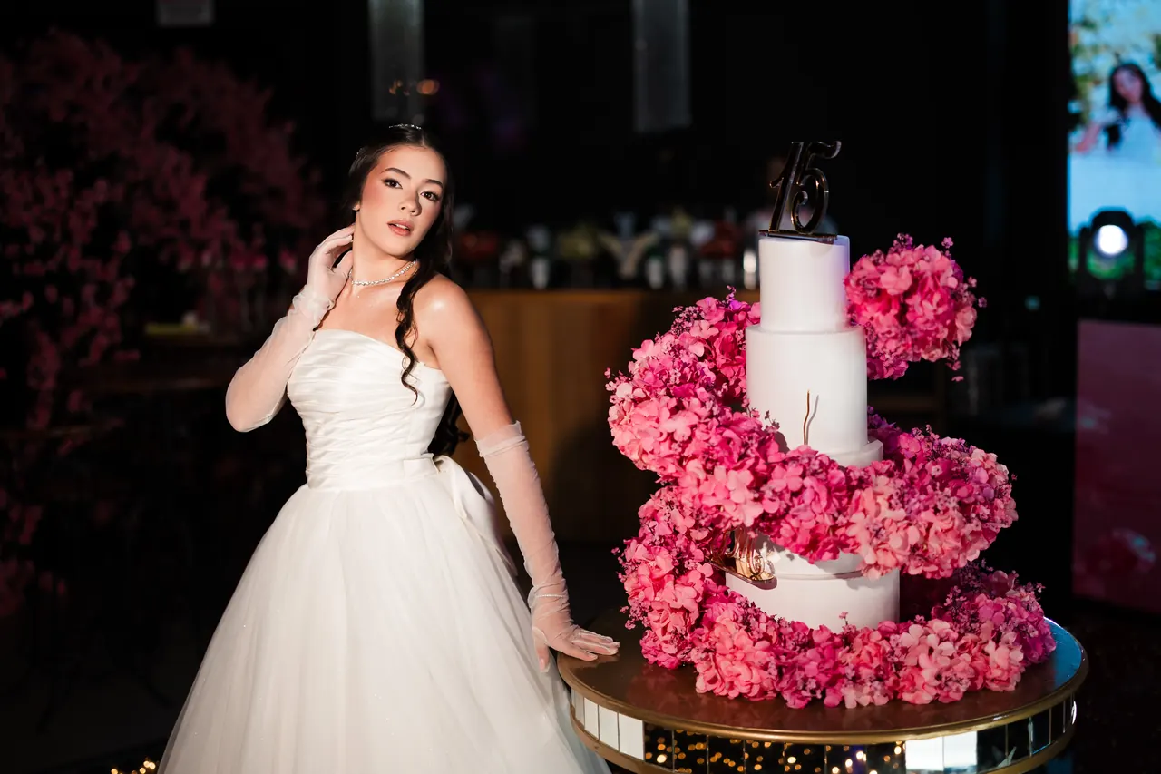 Debutante de vestido branco ao lado do bolo de 15 anos, com flores cor-de-rosa.