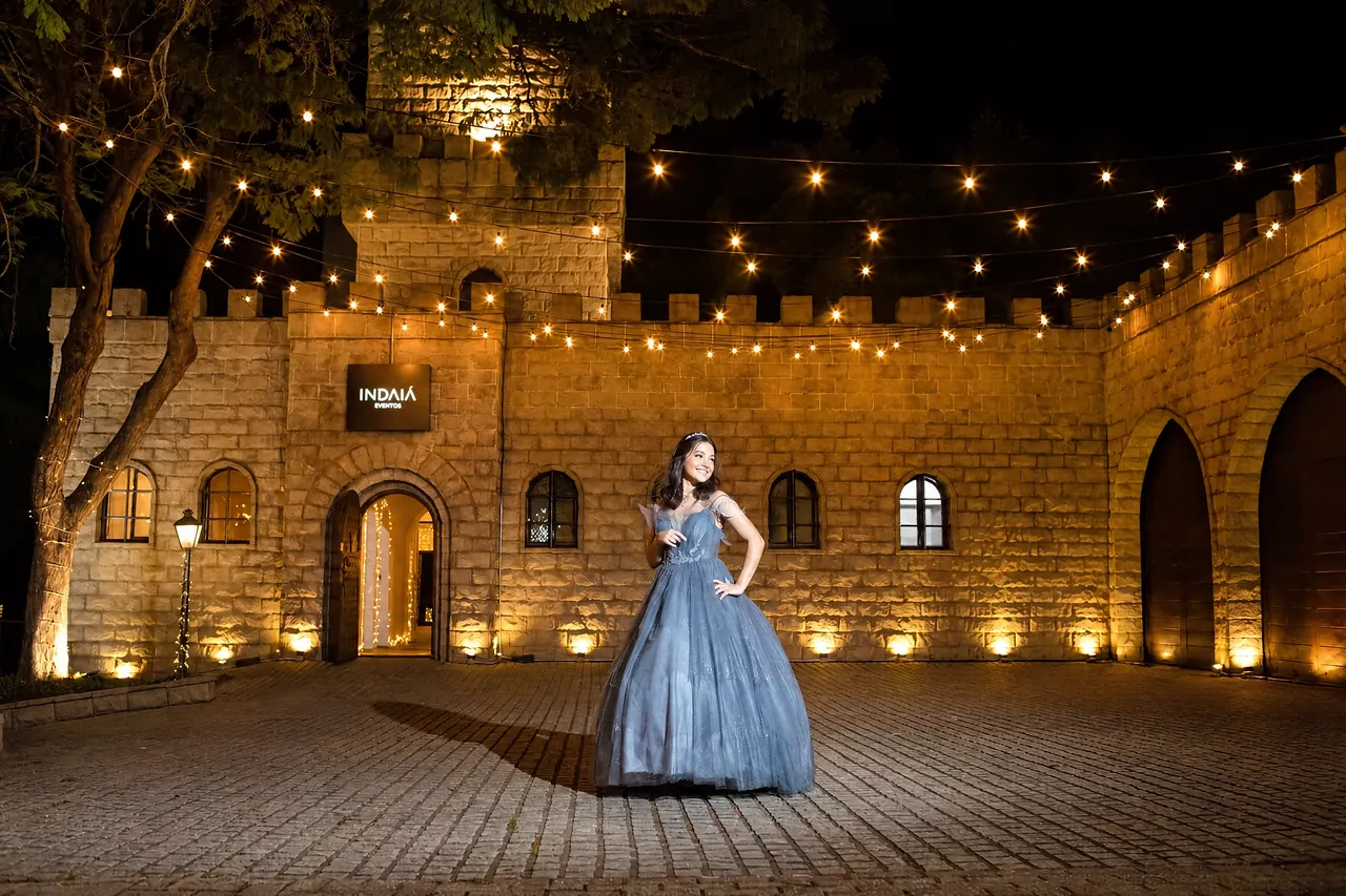 Debutante diante do Castelo Blumenau do Indaiá, com iluminação noturna e cordões de luzes sobre a fachada.