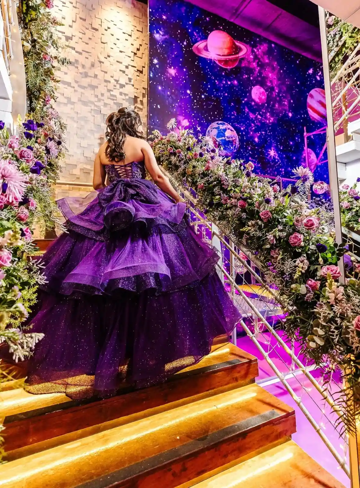 Debutante de vestido roxo em uma escadaria com flores e painel iluminado com planetas.