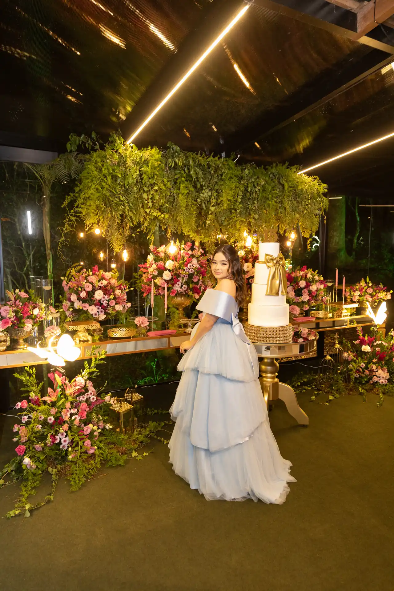 Debutante junto à mesa de bolo, flores e folhagens suspensas em uma celebração de Nova Veneza.