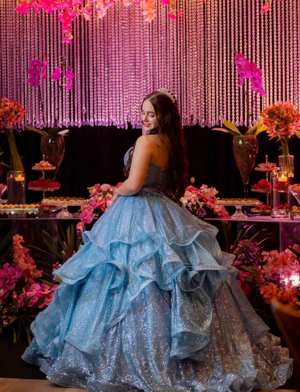 Debutante de vestido azul diante da mesa de doces, flores e cortina de cristais iluminada em rosa.